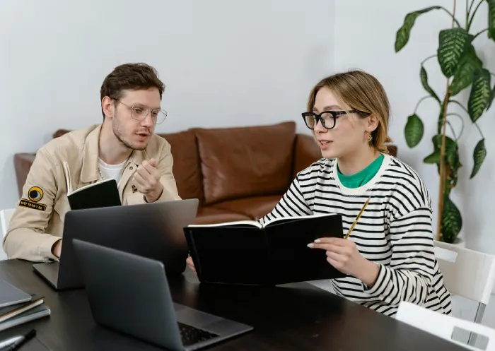 a man and woman talking in front of their macbook, holing notebooks